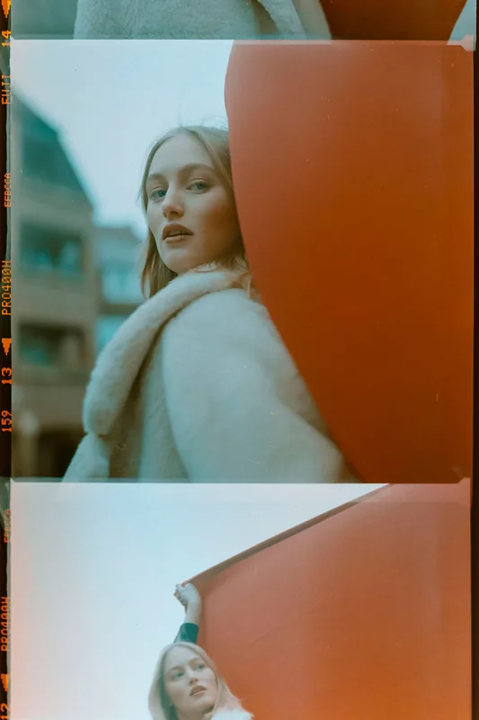 Analoges Portrait einer blonden Frau in einem weißen Pelzmantel hinter einer großen orangefarbenen Fläche.