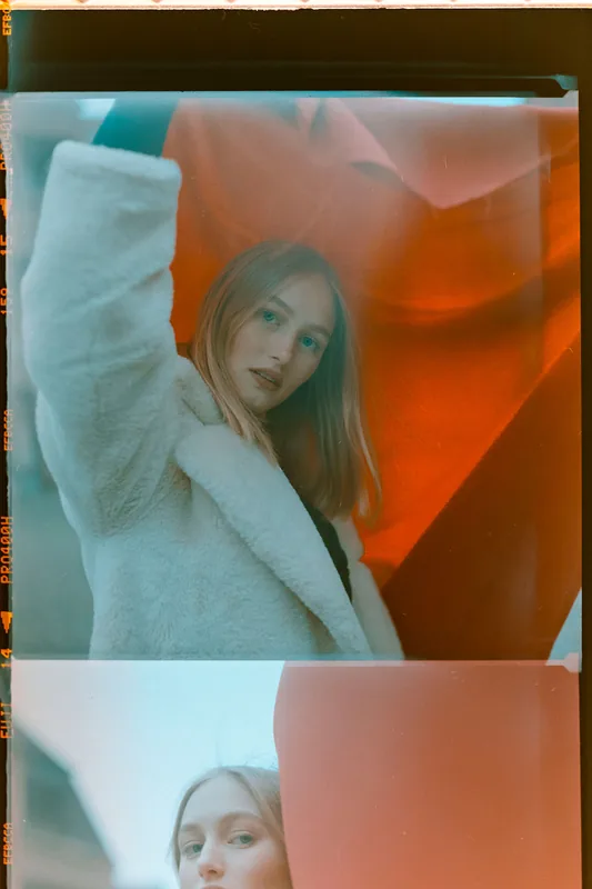 Analoges Portrait einer Frau mit blonden Haaren in einem weißen, flauschigen Mantel vor rotem Hintergrund.