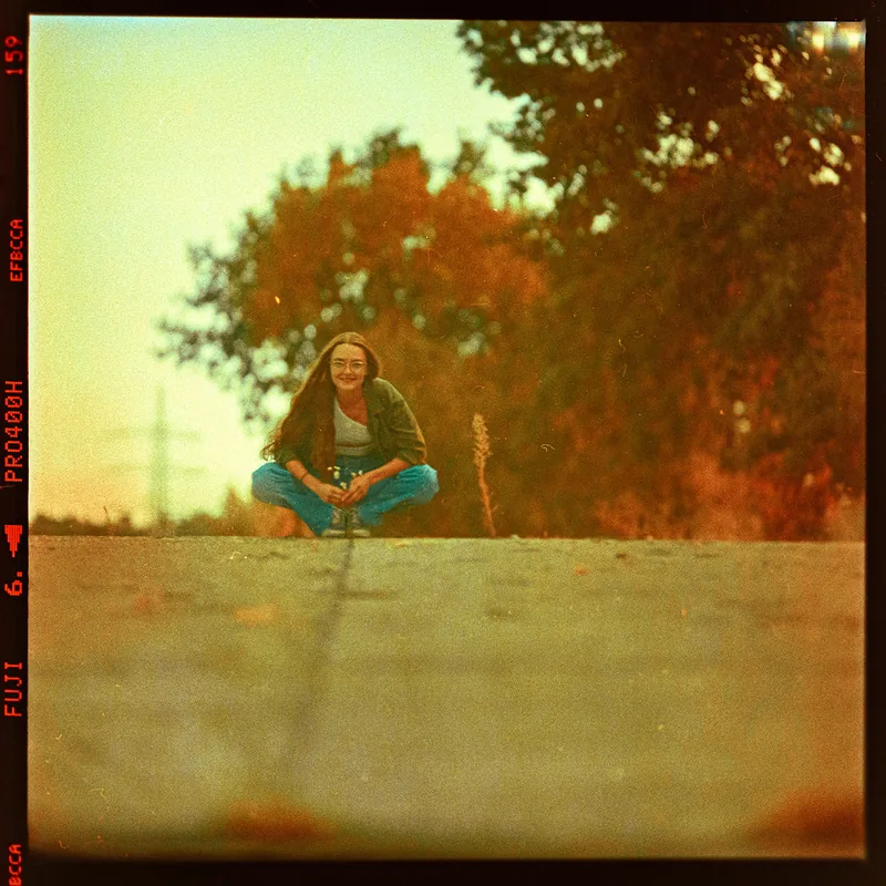 Analoges Portrait einer Frau mit Brille, die vor herbstlichen Bäumen hockt.