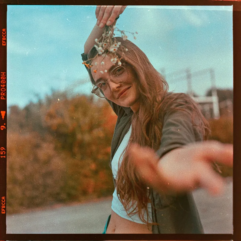 Analoges Portrait einer Frau mit Brille, die kleine Trockenblumen über ihrem Kopf hält.