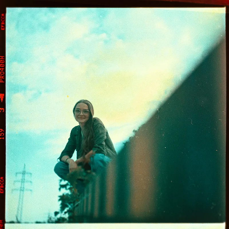 Analoges Portrait einer Frau mit Brille, die auf einem Vorsprung vor bewölktem Himmel sitzt.