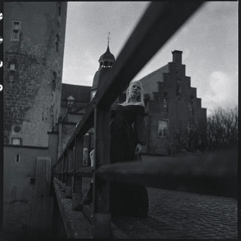 Analoges Portrait einer Frau, die an einer Brücke vor einem historischen Gebäude nach oben blickt.
