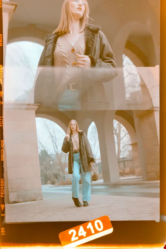 Analoges Portrait einer Frau in einem dunklen Mantel und heller Strickjacke unter steinernen Bögen.