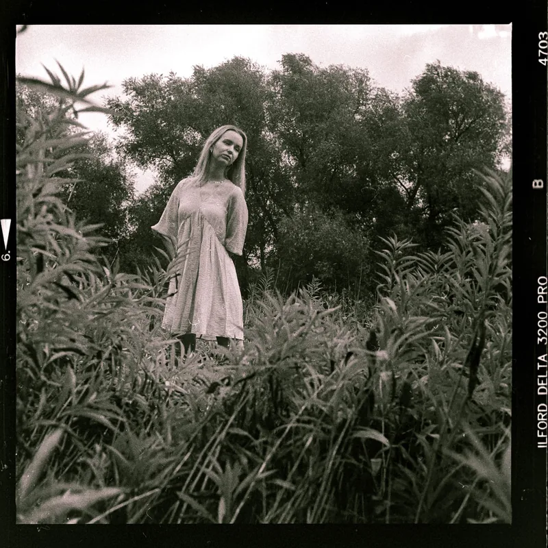 Analoges Portrait einer Frau in einem hellen Kleid inmitten von hohem Gras und Bäumen.