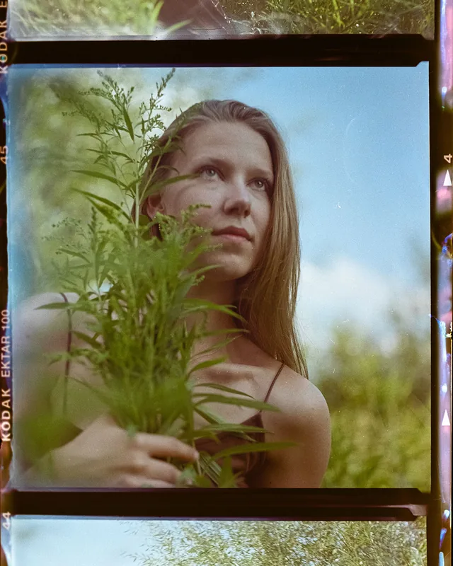 Analoges Portrait einer Frau hinter grünen Pflanzen vor blauem Himmel.
