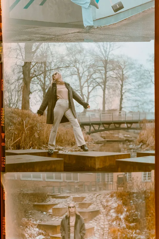 Eine Frau in hellem Outfit steht auf Betonblöcken vor einer winterlichen Parkkulisse im analogen Stil.