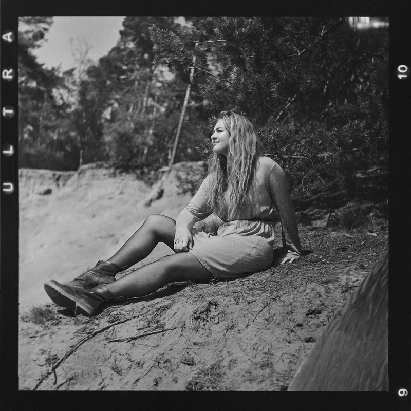 Analoges Portrait einer Frau mit langen Haaren, sitzend auf einer sandigen Böschung im Wald.