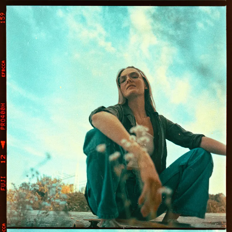 Analoges Portrait einer Frau mit Sonnenbrille und türkisfarbener Hose unter blauem Himmel.
