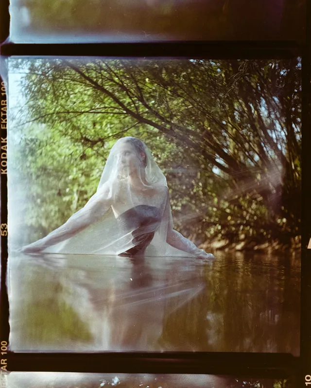 Analoges Portrait einer Frau mit weißem, transparentem Schleier im ruhigen Wasser zwischen grünen Bäumen.