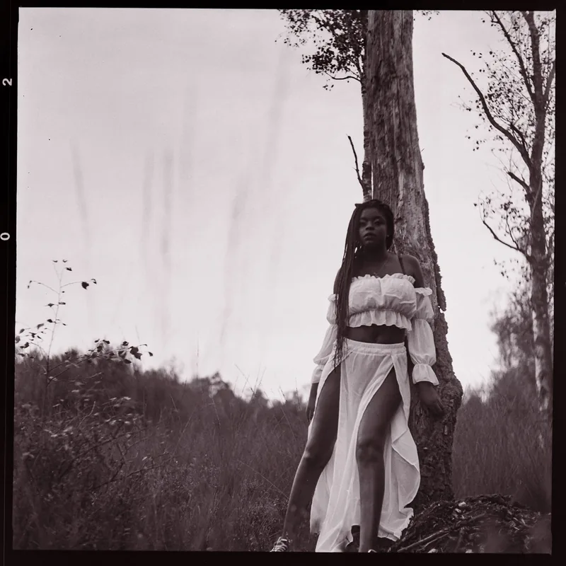 Analoges Portrait einer Frau in weißer Kleidung neben einem Baum im Freien.