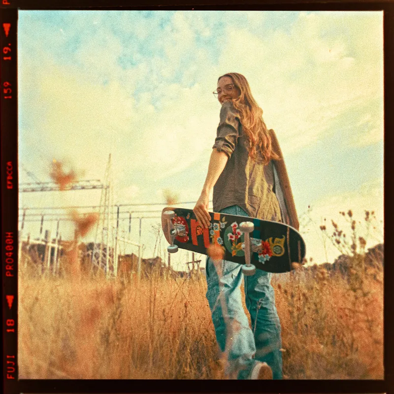 Analoges Portrait einer jungen Frau mit Brille und Skateboard in einem hohen Grasfeld.