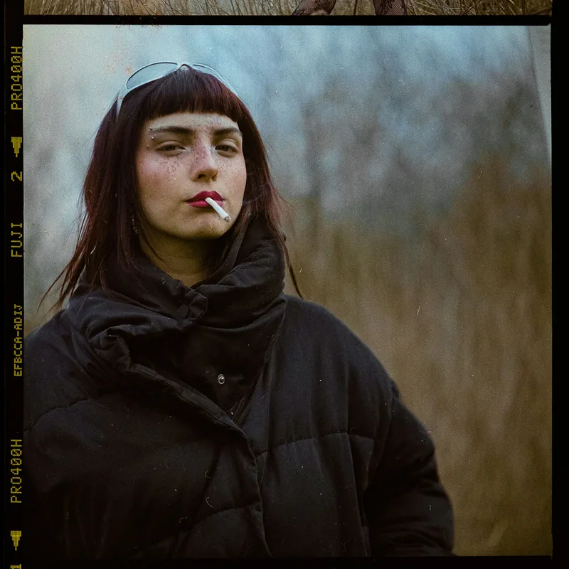 Analoges Portrait einer jungen Frau mit dunklem Pony, roten Lippen und einer Zigarette im Mund.