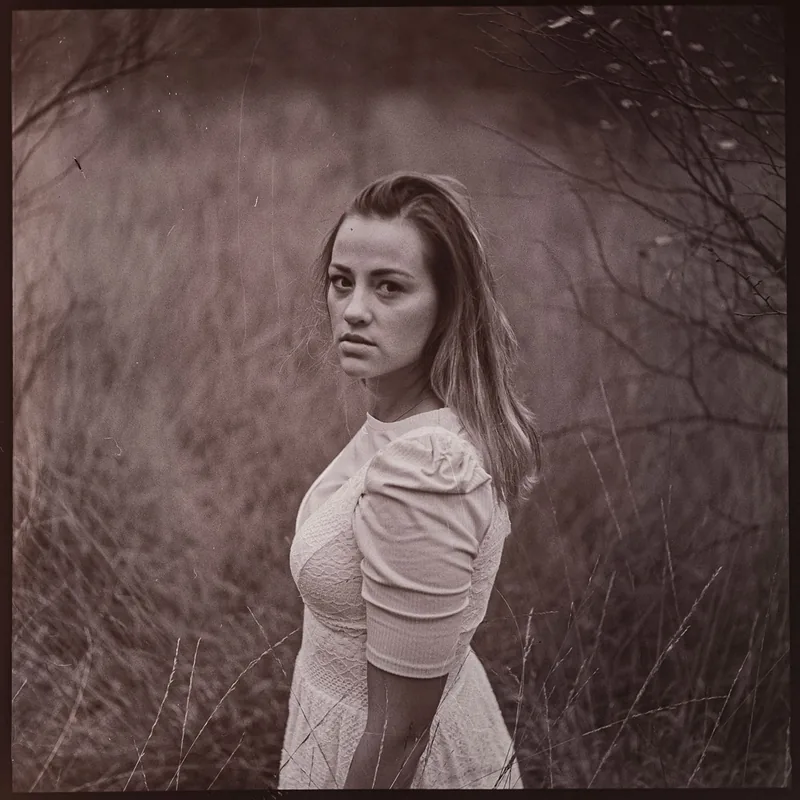 Analoges Portrait einer jungen Frau in einem hellen Kleid inmitten von trockenem Gras und Zweigen.