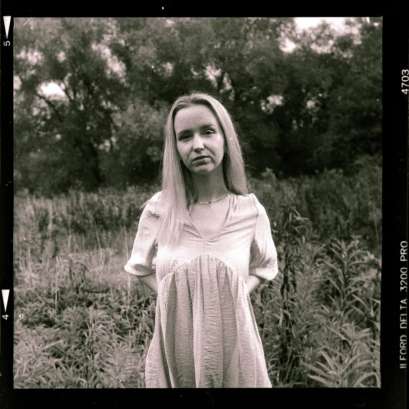 Analoges Portrait einer jungen Frau mit langen Haaren in einer Wiese vor Bäumen.