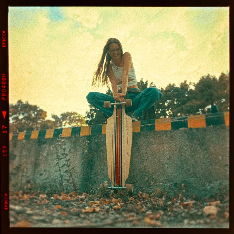 Analoges Portrait einer jungen Frau mit einem Longboard auf einer niedrigen Mauer.
