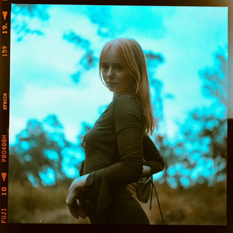 Analoges Portrait einer jungen Frau mit rötlichem Haar vor Bäumen und blauem Himmel.
