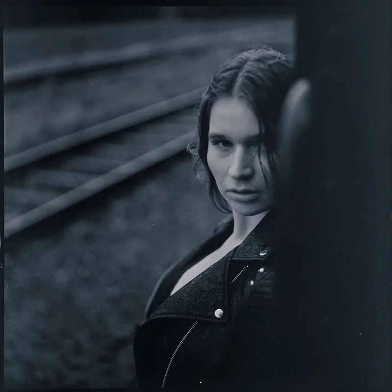 Analoges Portrait einer jungen Frau vor unscharfen Bahngleisen.