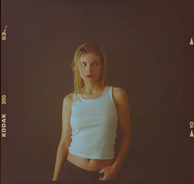 Analoges Portrait einer jungen Frau im weißen Tanktop vor dunklem Hintergrund.