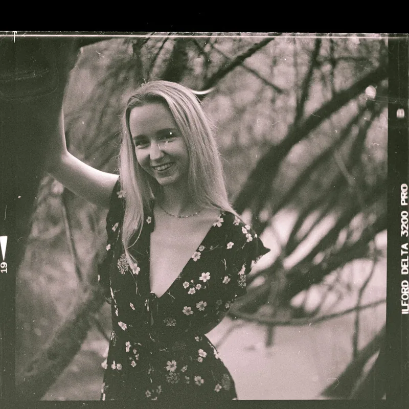 Analoges Portrait einer lächelnden Frau mit blondem Haar in einem floralen Kleid vor Bäumen.