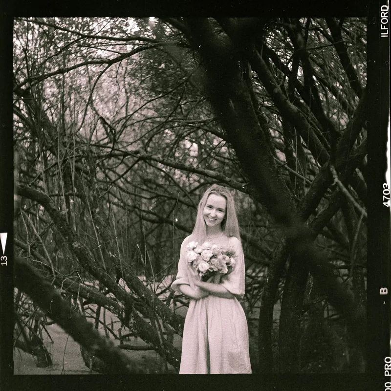 Analoges Portrait einer lächelnden Frau mit Blumenstrauß vor dichtem Geäst.