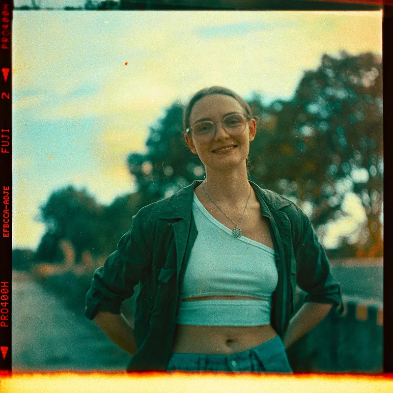 Analoges Portrait einer lächelnden jungen Frau mit Brille vor Bäumen.