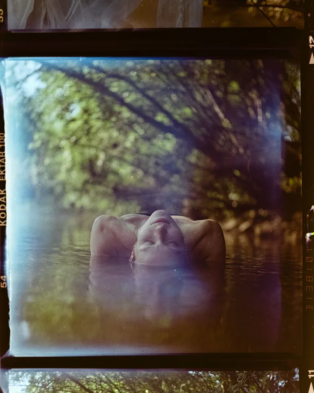 Analoges Portrait einer Person, die mit geschlossenen Augen im Wasser zwischen Bäumen treibt.