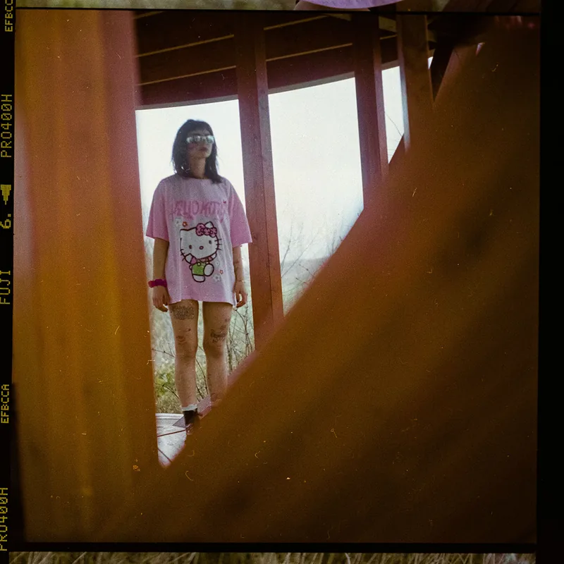 Analoges Portrait einer Person im pinken Hello-Kitty-Shirt unter einem Holzdach.