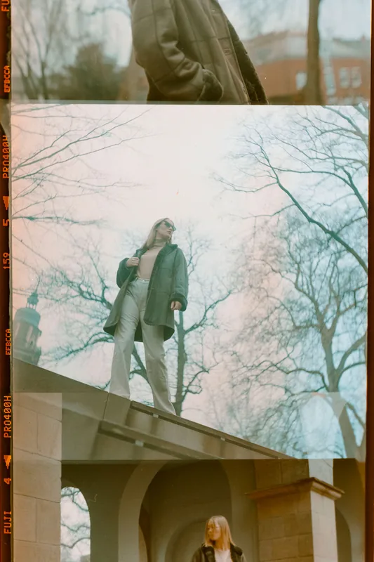 Analoges Portrait einer Person mit Sonnenbrille und heller Hose auf einer Steinmauer vor kahlen Bäumen.