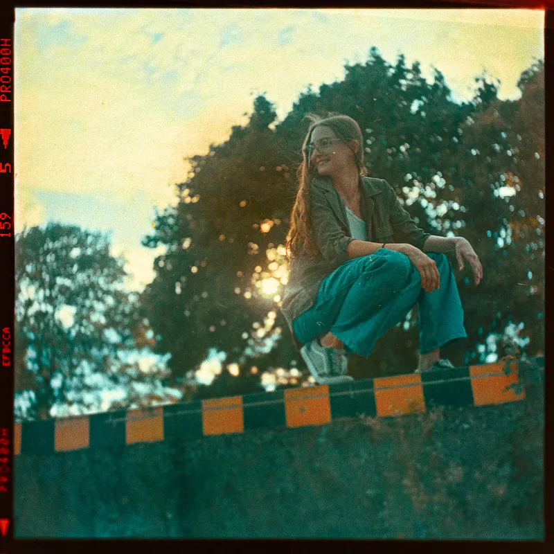Analoges Portrait einer sitzenden Frau vor Bäumen im warmen Gegenlicht der Abendsonne.