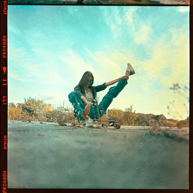 Analoges Portrait einer sitzenden Person mit Skateboard auf einer Betonmauer unter blauem Himmel.