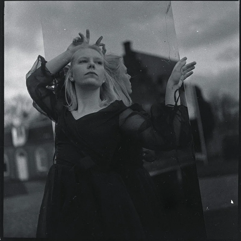 Analoges Schwarz-Weiß-Portrait einer blonden Frau mit ihrer Spiegelung in einer Glasscheibe.