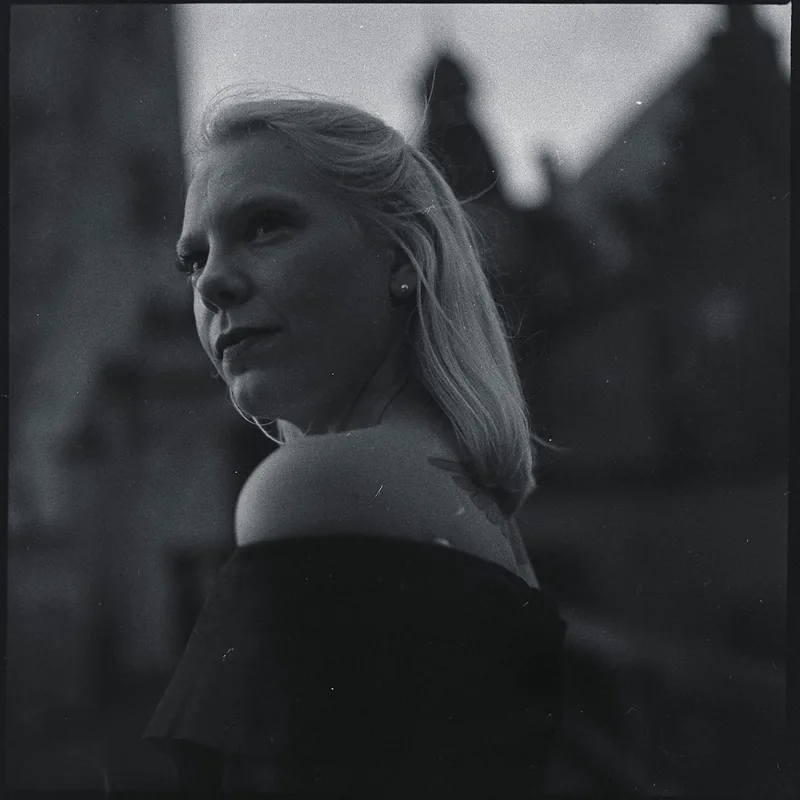 Analoges Schwarz-Weiß-Portrait einer blonden Frau vor unscharfen Gebäuden.