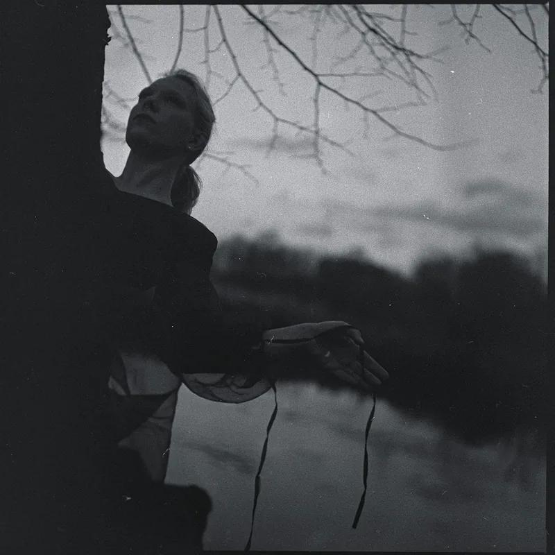 Analoges Schwarz-Weiß-Portrait einer Frau, die an einem Baum lehnt und mit ausgestreckter Hand dünne Bänder hält.