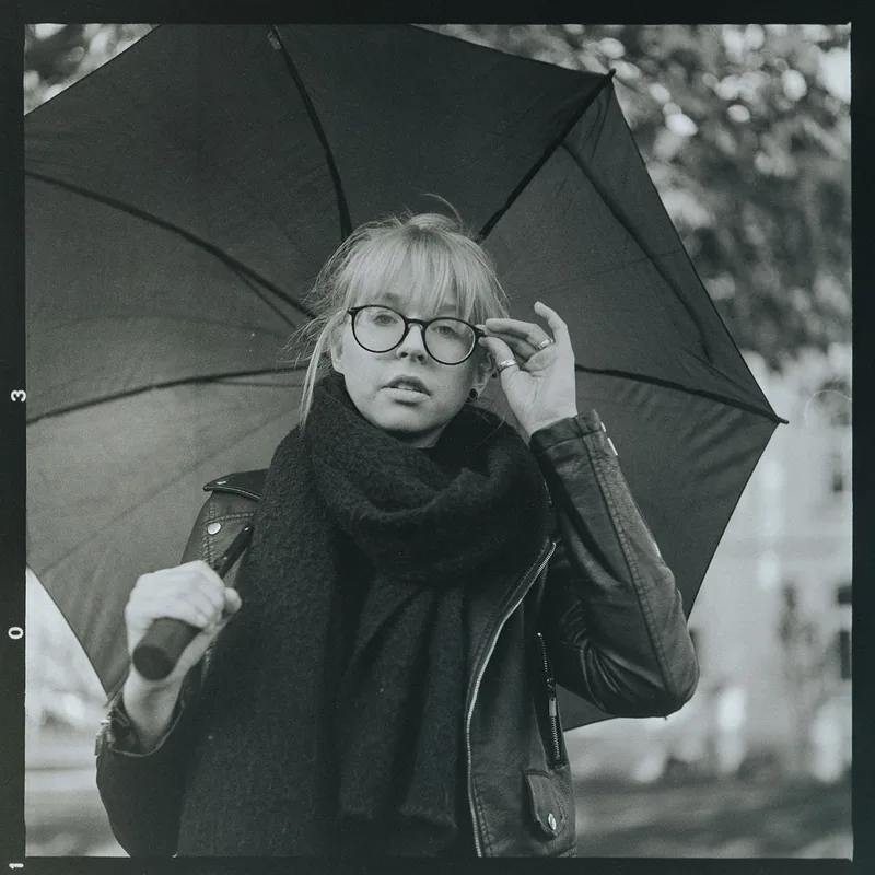 Analoges Schwarz-Weiß-Portrait einer Frau mit Brille und Schal unter einem großen Regenschirm.
