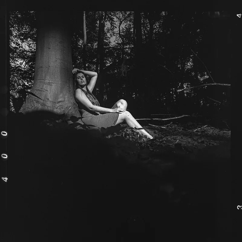 Analoges Schwarz-Weiß-Portrait einer Frau, die am Fuße eines großen Baumes im Wald lehnt.