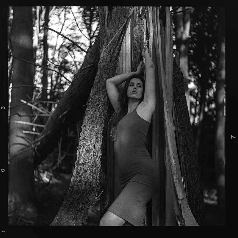Analoges Schwarz-Weiß-Portrait einer Frau, die sich an einen aufgesplitterten Baumstamm im Wald lehnt.