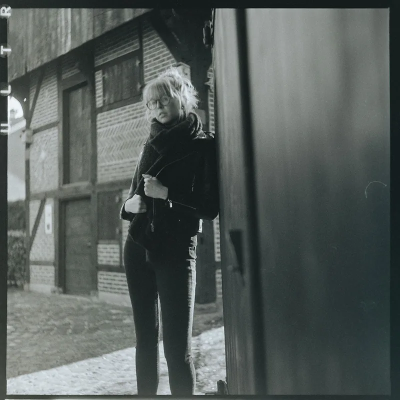 Analoges Schwarz-Weiß-Portrait einer jungen Frau mit Brille vor einem Fachwerkhaus.