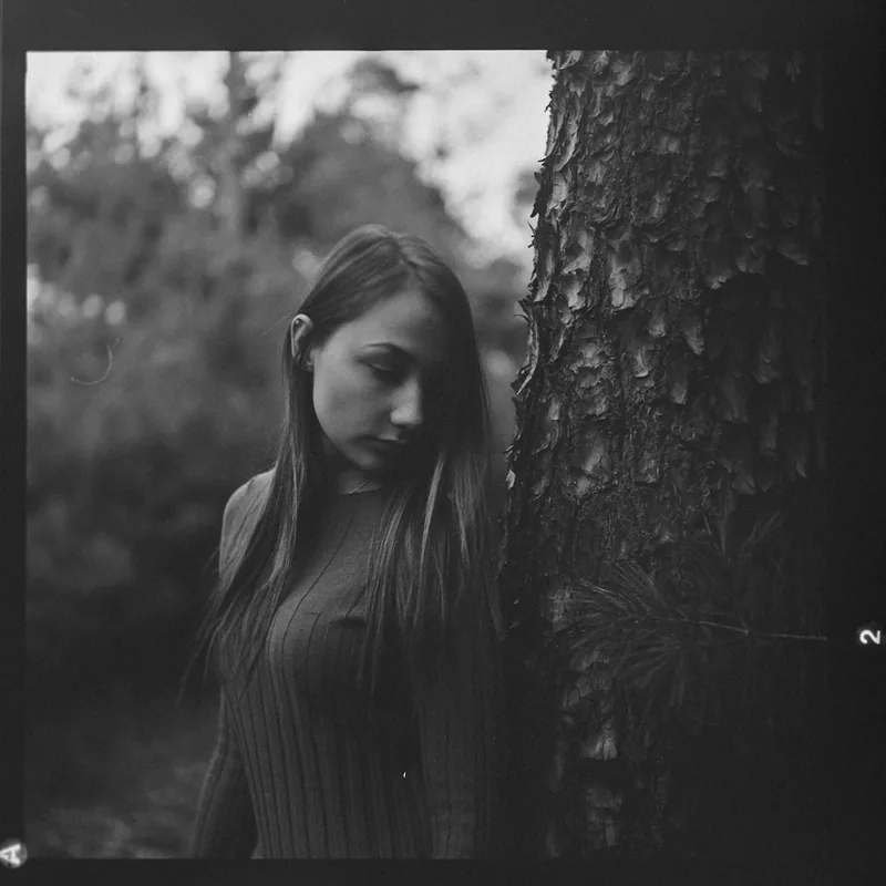 Analoges Schwarz-Weiß-Portrait einer jungen Frau neben einem Baumstamm im Wald.