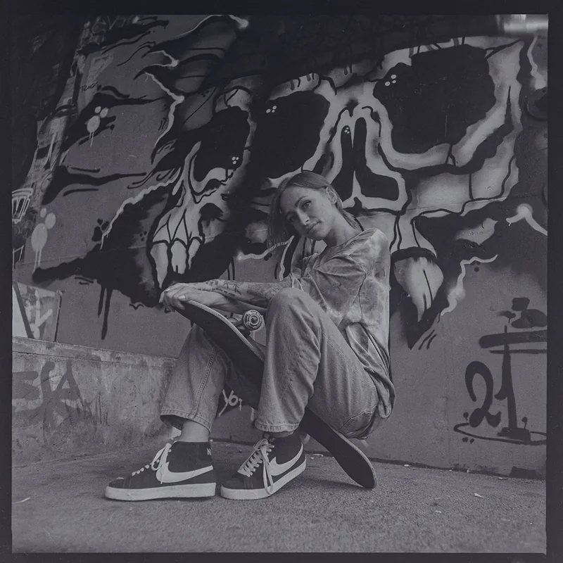 Analoges Schwarz-Weiß-Portrait einer jungen Frau, die auf einem Skateboard vor einer Graffiti-Wand sitzt.