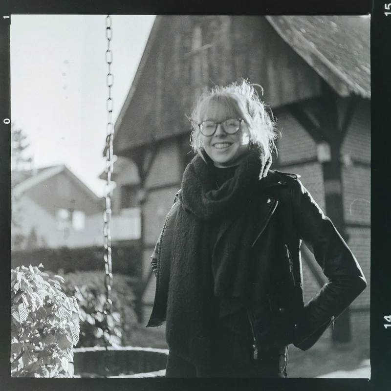 Analoges Schwarz-Weiß-Portrait einer lächelnden Frau mit Brille und Schal vor einem Holzgebäude.
