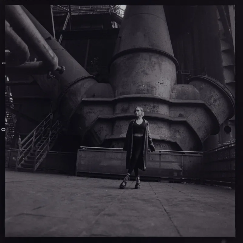 Analoges Schwarzweiß-Portrait einer Frau vor massiven industriellen Rohrleitungen.