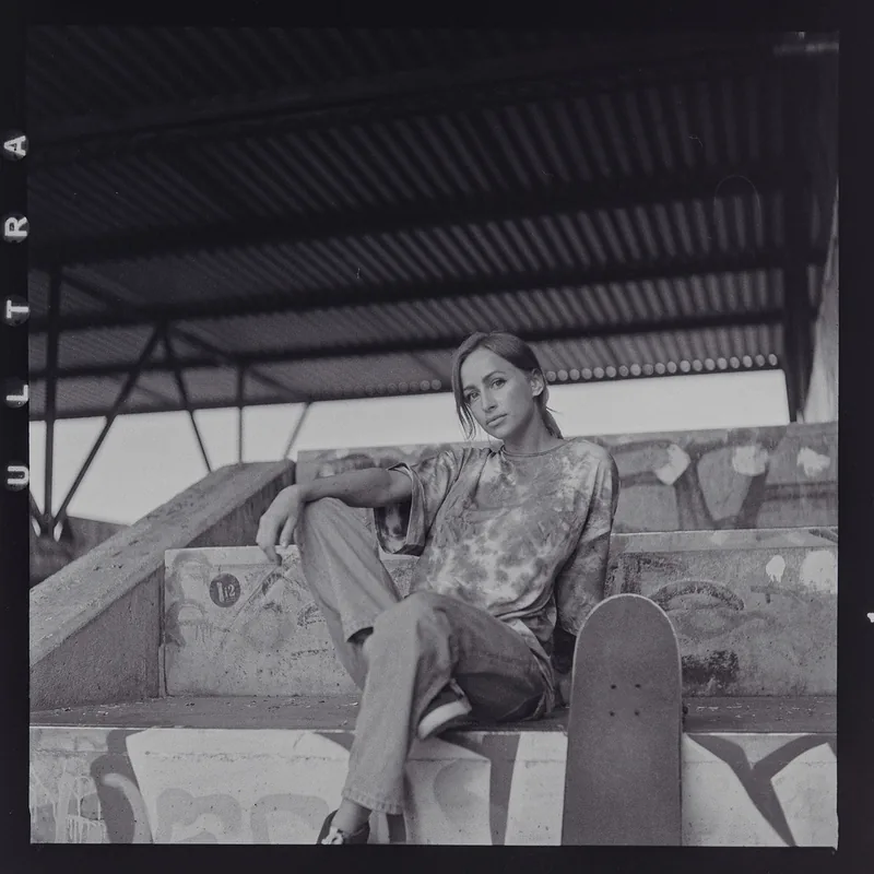 Analoges Schwarzweiß-Portrait einer Frau, die neben einem Skateboard auf Betonstufen sitzt.