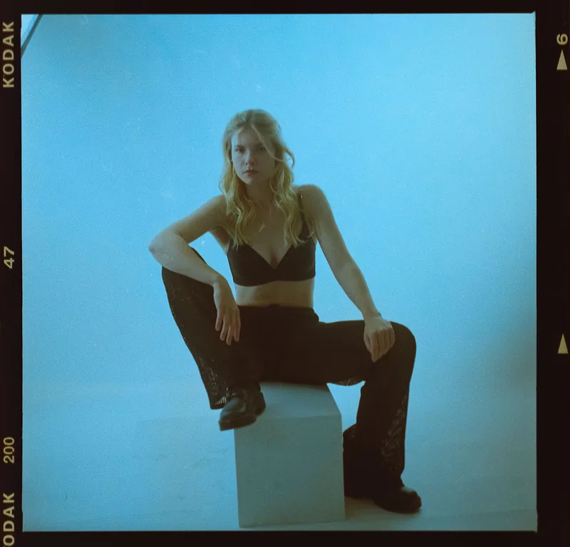 Analoges Studioportrait einer Frau in schwarzer Kleidung, sitzend auf einem weißen Block vor blauem Hintergrund.