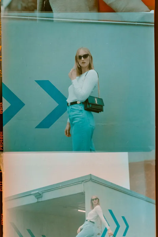 Blonde Frau mit Sonnenbrille, weißem Oberteil und blauer Jeans vor blaugrünem Hintergrund mit Pfeil-Motiv.