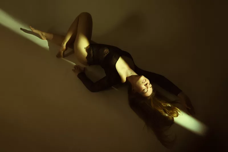 Boudoirfoto einer Frau in schwarzem Bodysuit unter einem hellen Lichtstrahl.