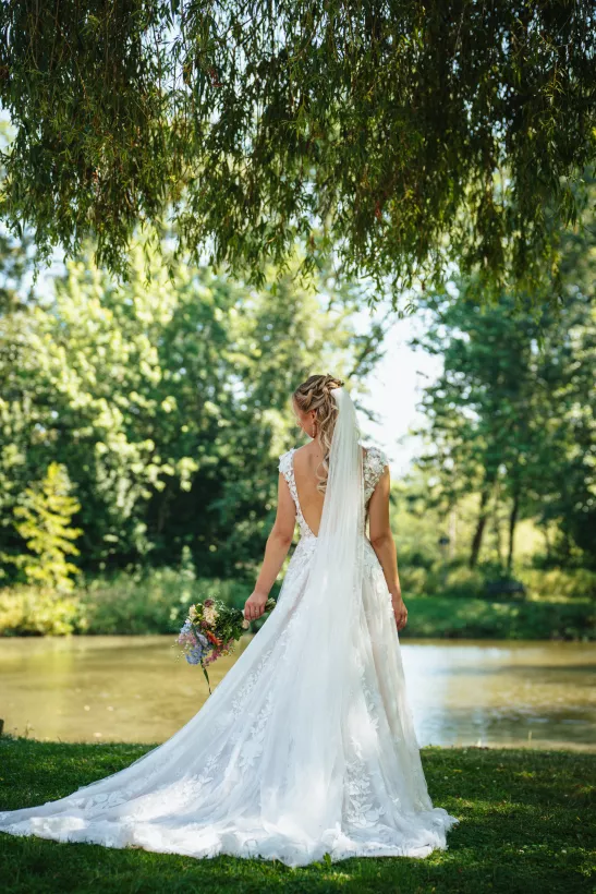 Braut im weißen Hochzeitskleid mit Schleier und Bouquet vor einem Teich unter Weidenzweigen