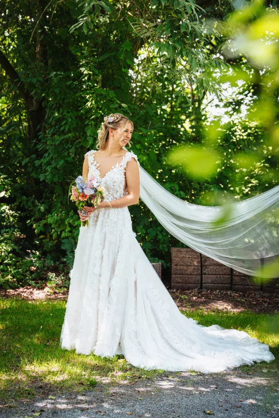 Braut in weißem Hochzeitskleid mit Blumenstrauß und langem Schleier vor grüner Natur