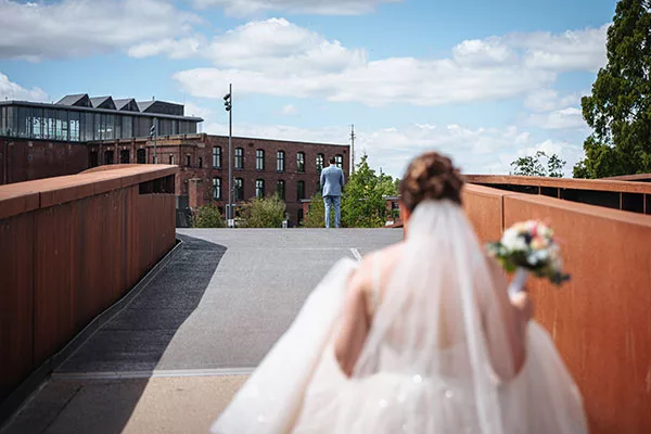 Braut mit Schleier und Bouquet, die von einem Steg aus auf den Bräutigam vor einem Backsteingebäude blickt.