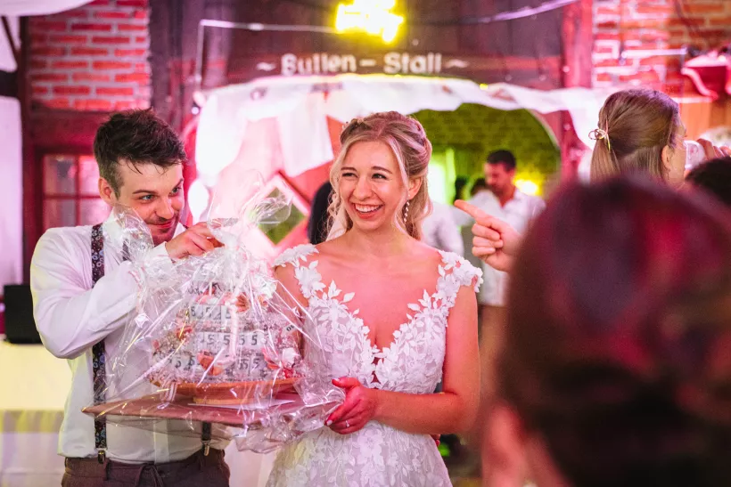 Lachende Braut und Mann mit einer in Folie verpackten Hochzeitstorte bei einer Hochzeitsfeier.