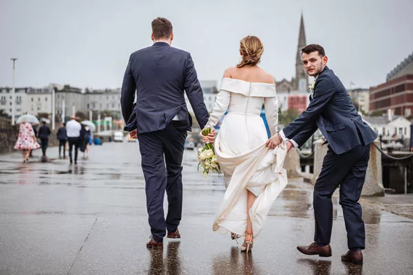Braut im weißen Hochzeitskleid und zwei Männer in Anzügen gehen über eine nasse Promenade.